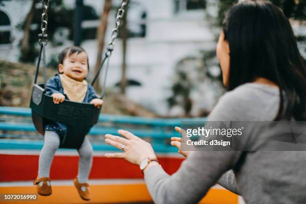 mother and baby girl having fun time in the park while mother swinging baby girl on swing - baby swing stock pictures, royalty-free photos & images