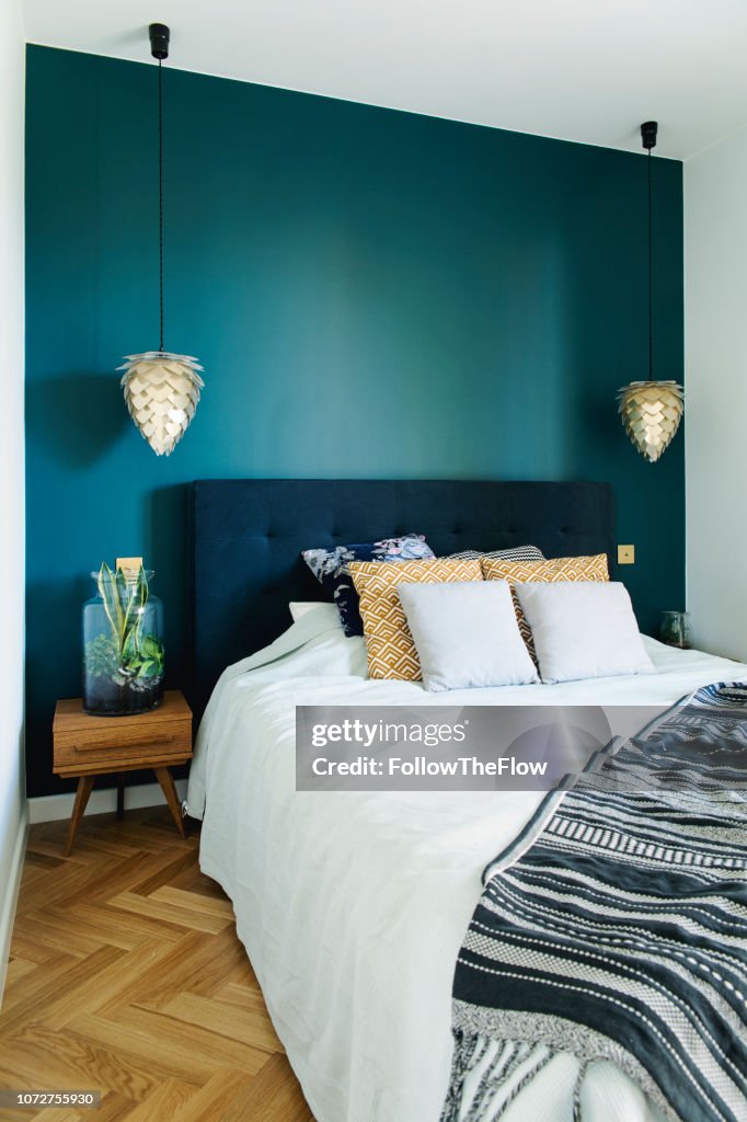 Stylish and modern sunny bedroom with small wooden table , white bedding and colours pilows. Space with blue walls and brown wooden parquet.