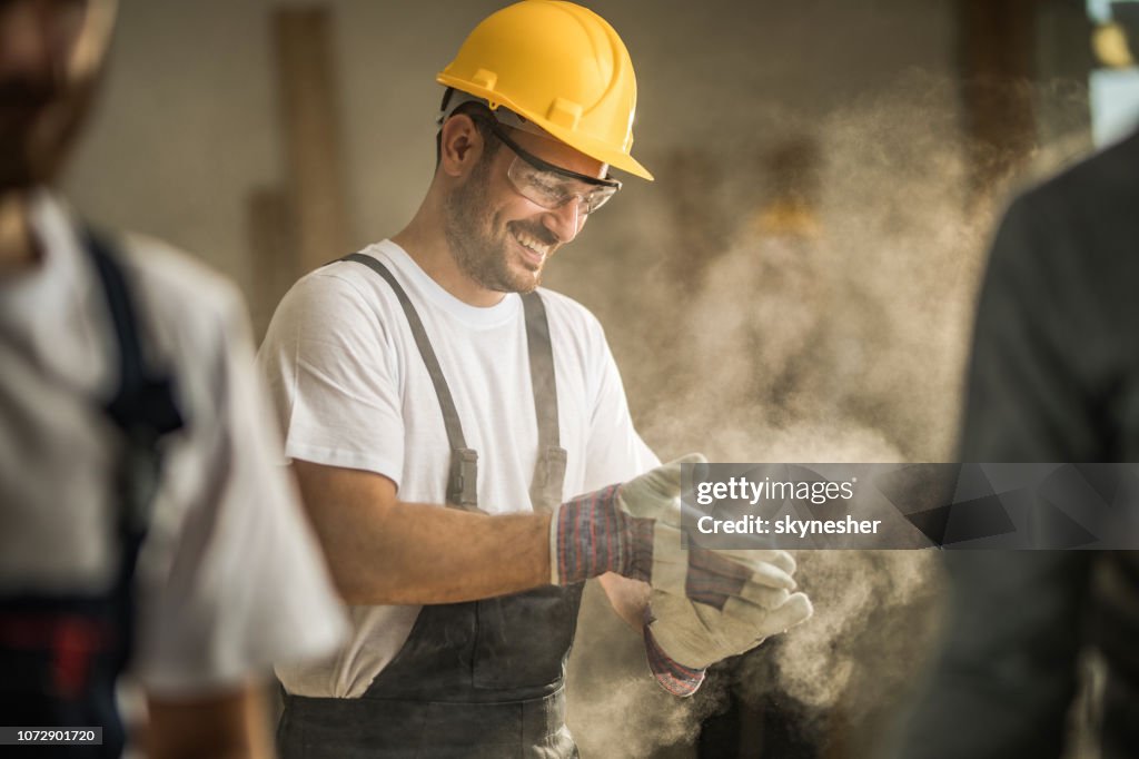 Feliz trabajador manual limpieza sus guantes de serrín en el sitio de construcción.