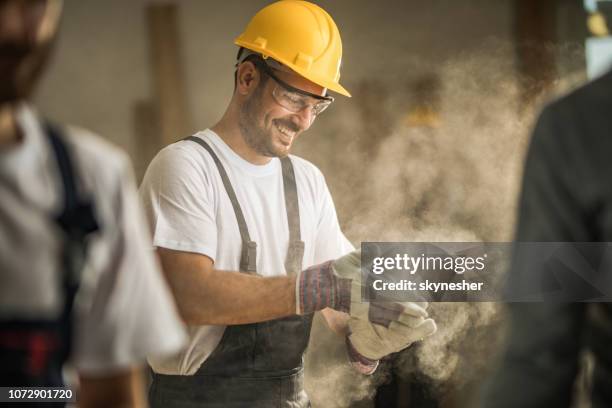 happy manual worker cleaning his gloves from sawdust at construction site. - industrial cleaning stock pictures, royalty-free photos & images