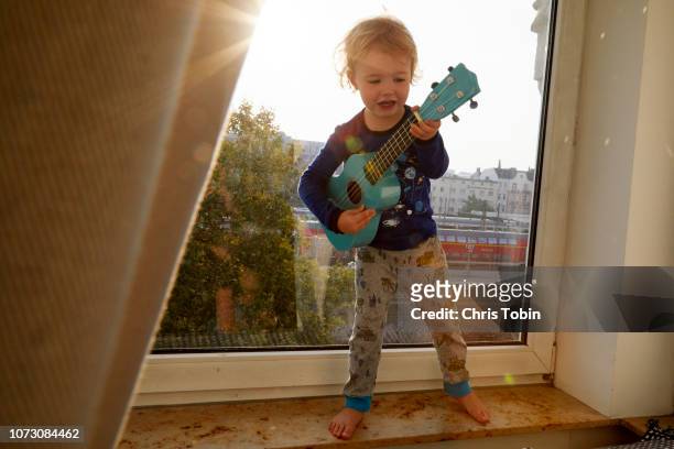 toddler playing ukulele standing on windowsill with sun flare - rock music stock-fotos und bilder