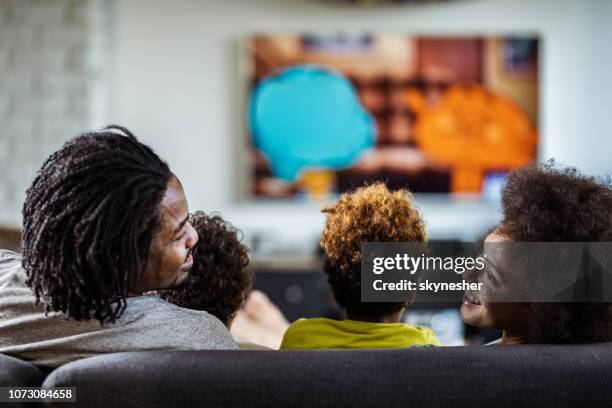 vue arrière du heureux parents noirs parler en regardant la télévision avec leurs enfants à la maison. - black people watching tv photos et images de collection
