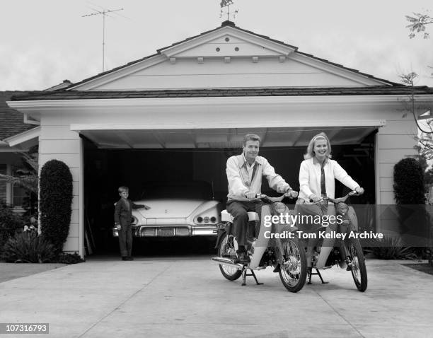Couple smiles while sitting on their Honda 50cc scooters, on their driveway, in front of their garage, 1958. Their son stands in the background,...