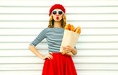 Portrait beautiful young woman blowing lips making air kiss wearing red beret holding in hands paper bag with long white bread baguette on white wall background