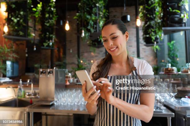 américa latina camarera alegre tomando un descanso chateando en smartphone sonriendo mientras está parado detrás de barra de bar - happy merchant fotografías e imágenes de stock