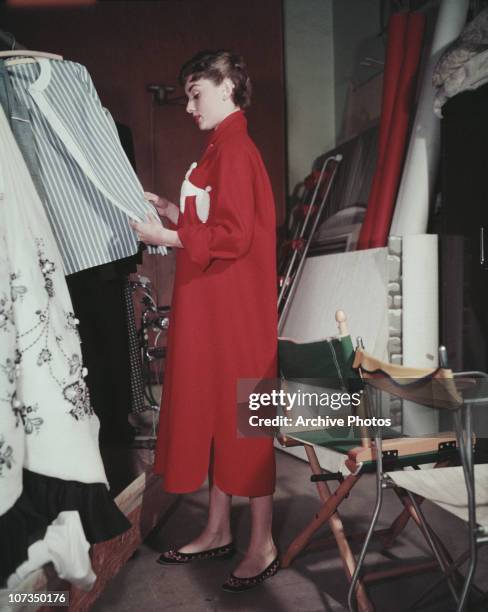 Belgian-born actress Audrey Hepburn browsing through the wardrobe at a photographer's studio, circa 1955. She is wearing a full length red coat dress...