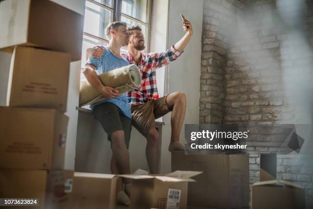 happy embraced gay couple taking a selfie while relocating into new apartment. - gay couple new house stock pictures, royalty-free photos & images