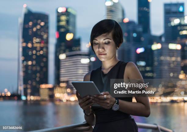 business woman using digital tablet in front of singapore skyline, at night - singapore cityscape stock pictures, royalty-free photos & images