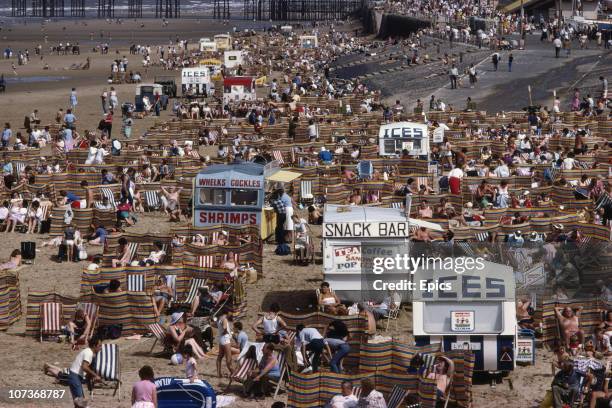 Crowds of holidaymakers and kiosks offering tea, coffee, ices, whelks and cockles and shrimps as snacks on the beach in the seaside resort of...