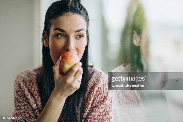 pensive woman eating an apple by the window. - apple bite out stock pictures, royalty-free photos & images