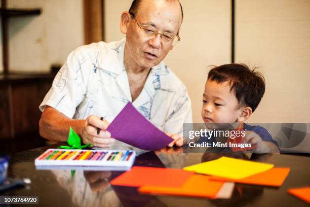 japanese man and little boy sitting at a table, making origami animals using brightly coloured paper. - origami asia stock pictures, royalty-free photos & images