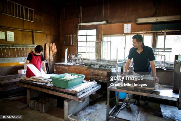 two people, man and woman making traditional washi paper. trays of pulp and wooden frames and drying racks. - 手漉きの紙 ストックフォトと画像