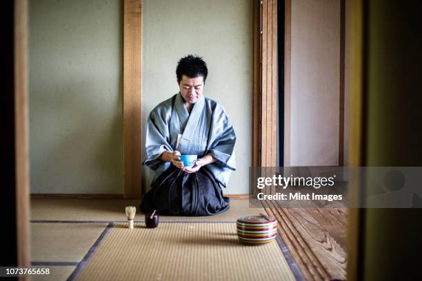 japanese man wearing traditional kimono kneeling on tatami mat, holding tea bowl, during tea ceremony. - tea ceremony stock pictures, royalty-free photos & images