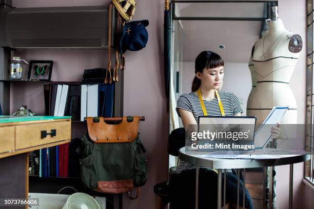 japanese female fashion designer working in her studio, sitting at table, looking at fabric samples. - design laptop woman stock-fotos und bilder