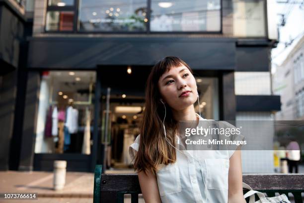 japanese woman with long brown hair wearing white short-sleeved blouse sitting on a bench, wearing earphones, listening to music. - magasin musique photos et images de collection