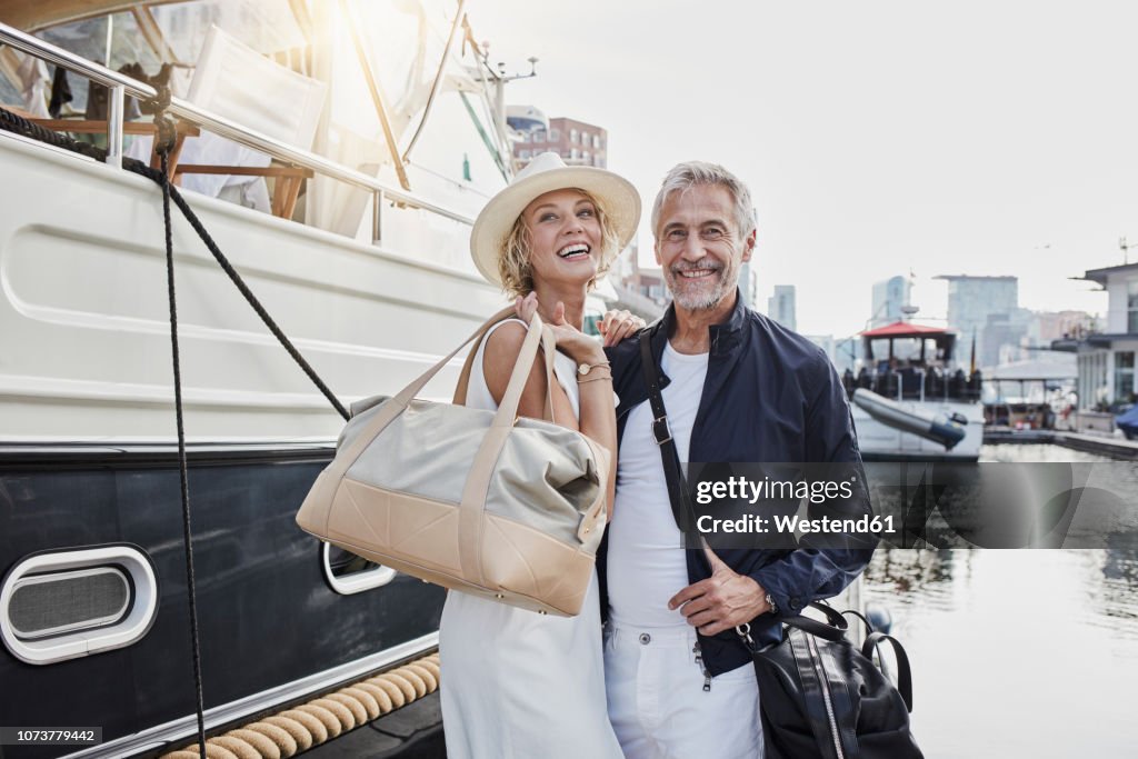 Older man and young woman standing with travelling bags on jetty next to yacht