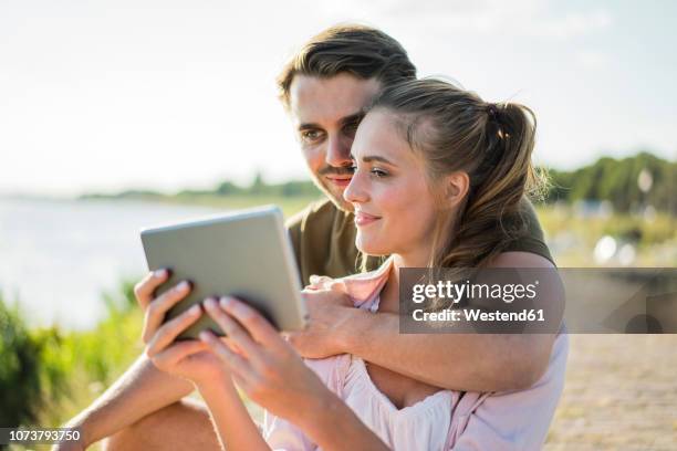 smiling couple at the riverside in summer holding tablet - tablet paar sommer stock-fotos und bilder