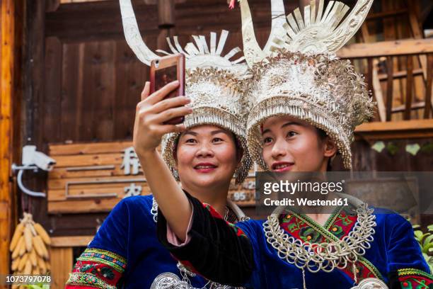 china, guizhou, two miao women wearing traditional dresses and headdresses taking a selfie with smartphone - tribal stock pictures, royalty-free photos & images