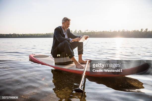 businessman sitting on paddleboard on a lake using tablet and earphones - barefoot men stock-fotos und bilder