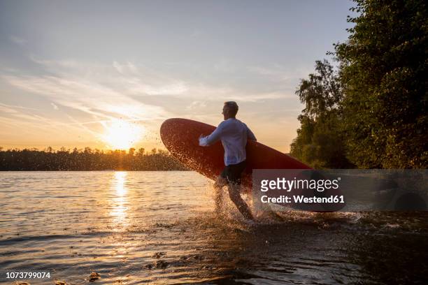 man with paddleboard walking into lake by sunset - 浮き板 ストックフォトと画像
