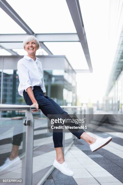 happy senior woman sitting on railing in the city looking around - geländer stock-fotos und bilder