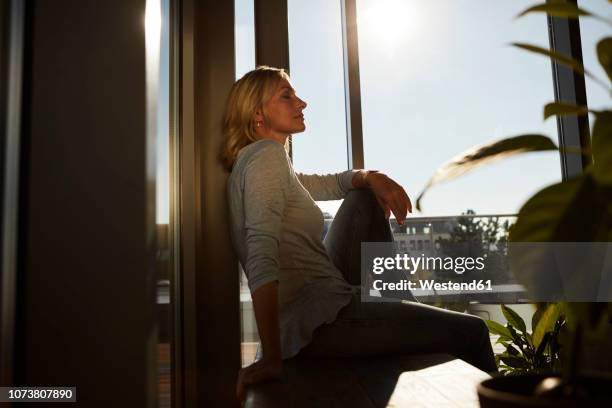 relaxed mature woman sitting in sunlight at home - window sunlight stock-fotos und bilder