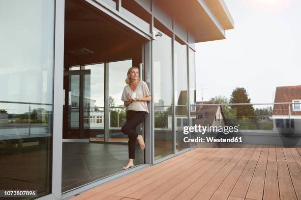 smiling mature woman with cup of coffee standing at roof terrace at home - balcony window stock pictures, royalty-free photos & images