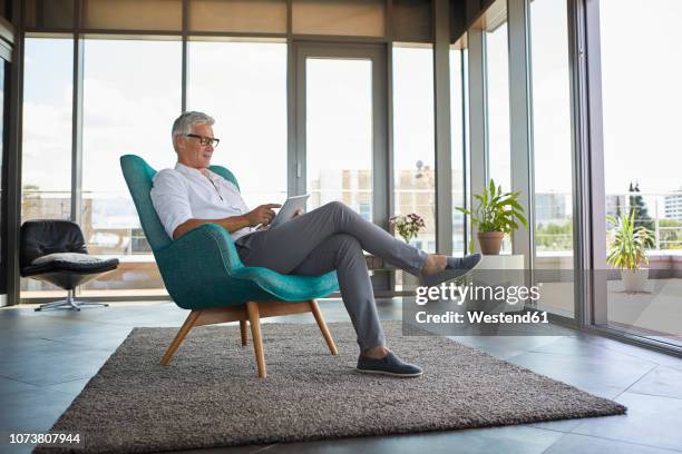 mature man sitting in armchair at home using tablet - mann ganzkörper casual stock-fotos und bilder