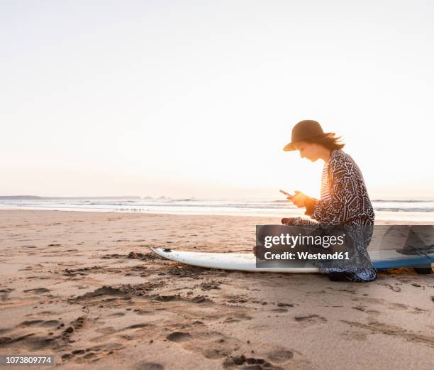 young woman sitting on surfboard at the beach, using smartphone - surfboard stock pictures, royalty-free photos & images