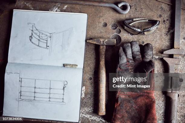 still life of artisan metalworker's tools, sketches in a notebook and a blackened thick protective glove - workbench stock pictures, royalty-free photos & images