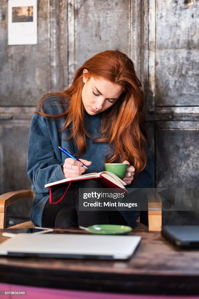 Young woman with long red hair sitting at table, holding notebook and cup of coffee.