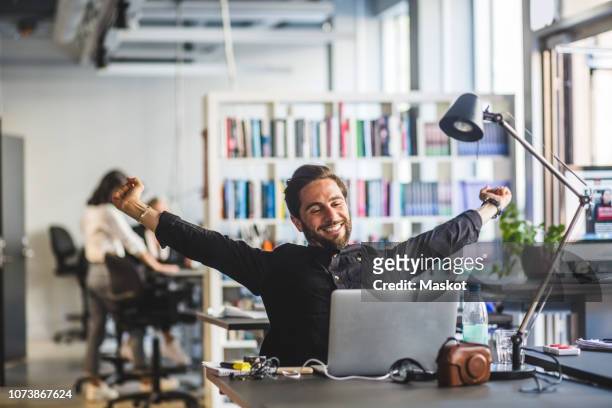 businessman sitting with arms outstretched at desk in office - dehnen stock-fotos und bilder