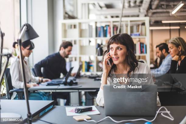 businesswoman looking away while talking on mobile phone at desk in creative office - mobile worker stockfoto's en -beelden