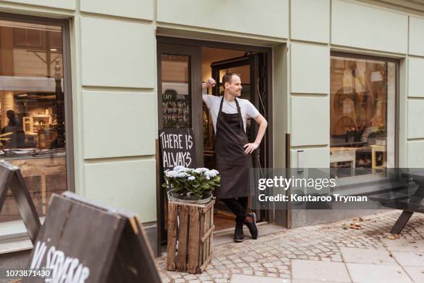 full length of salesman standing at doorway of cafe - boutique entrance stock pictures, royalty-free photos & images