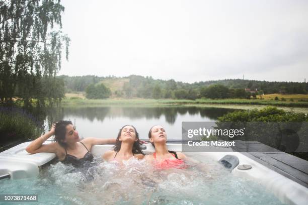 female friends enjoying in hot tub with eyes closed against lake during weekend getaway - hot tub party stock pictures, royalty-free photos & images