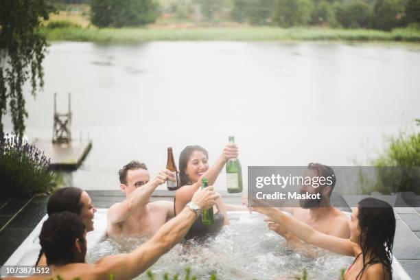 carefree male and female friends enjoying drinks in hot tub against lake during weekend getaway - hot tub party stock pictures, royalty-free photos & images