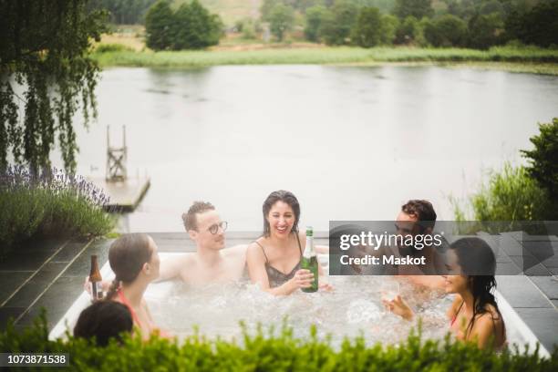 cheerful male and female friends enjoying drinks in hot tub against lake during weekend getaway - hot tub party stock pictures, royalty-free photos & images