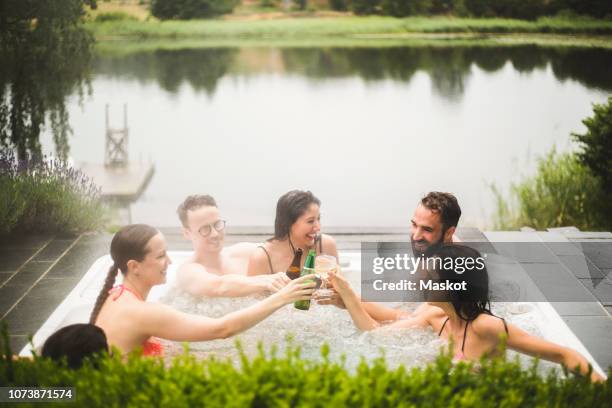 cheerful male and female friends toasting drinks in hot tub against lake during weekend getaway - hot tub party stock pictures, royalty-free photos & images