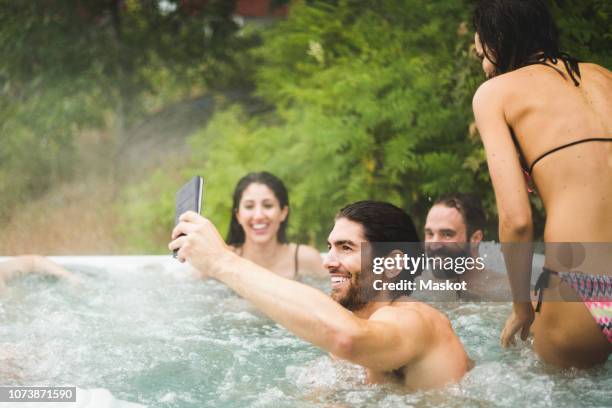 young man taking selfie through mobile phone while friends enjoying in hot tub during weekend getaway - hot tub party stock pictures, royalty-free photos & images