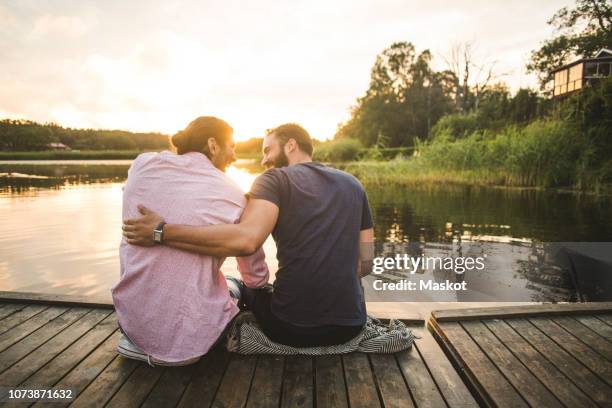 rear view of smiling male friends sitting on jetty over lake during sunset - male friendship stock pictures, royalty-free photos & images