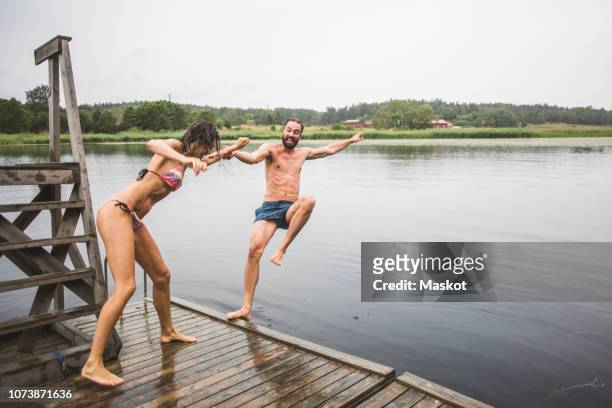 full length of playful woman pushing male friend in lake while standing on jetty during weekend getaway - stockholm summer stockfoto's en -beelden