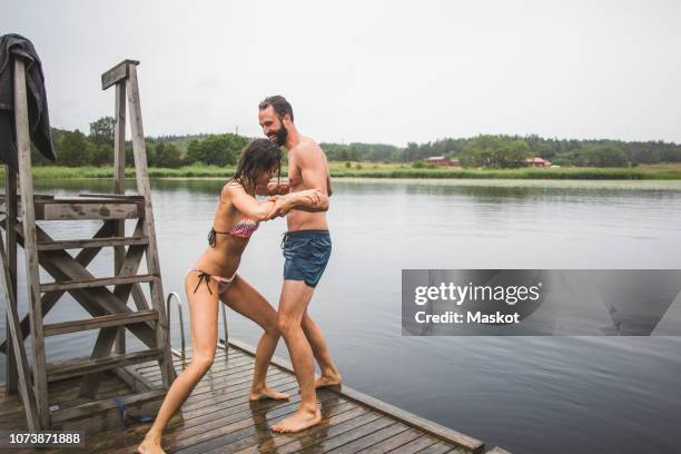 playful woman pushing male friend in lake while standing on jetty during weekend getaway - stockholm summer stock-fotos und bilder