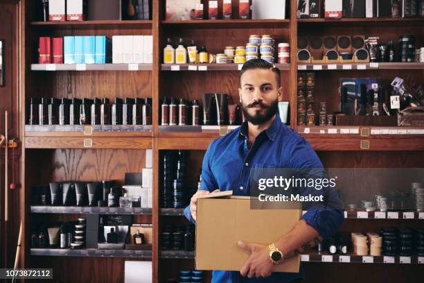 portrait of confident salesman carrying box while standing against rack in deli - cosmetics box stock pictures, royalty-free photos & images