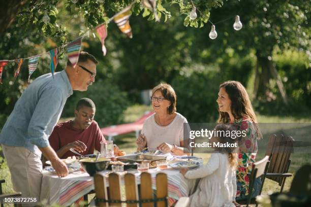 senior man and granddaughter talking while having lunch with family in backyard during party - outdoor lunch stock pictures, royalty-free photos & images