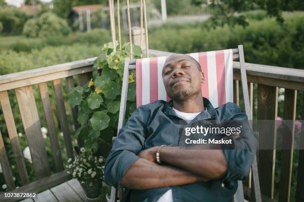 mid adult man with closed eyes relaxing on deck chair at porch - deckchair stock pictures, royalty-free photos & images