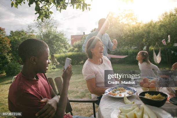 boy photographing cheerful family enjoying lunch at table during garden party - photographing garden stock pictures, royalty-free photos & images