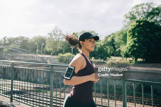 confident sportswoman listening music through in-ear headphones while jogging on bridge in city - jogging stock-fotos und bilder