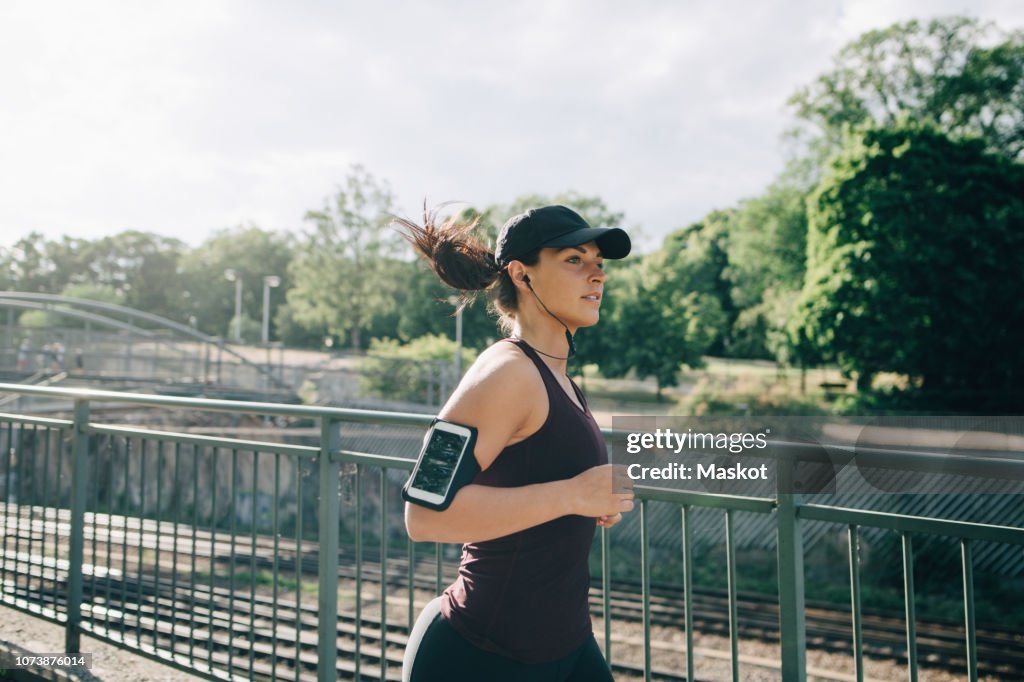 Confident sportswoman listening music through in-ear headphones while jogging on bridge in city