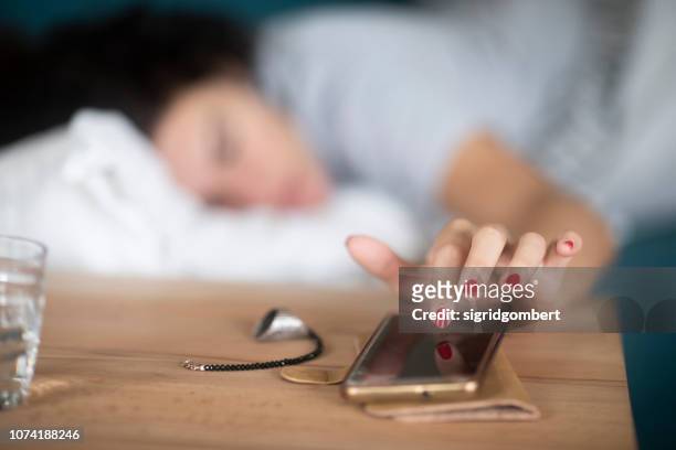 woman in bed turning off the alarm on her mobile phone - dormitar fotografías e imágenes de stock