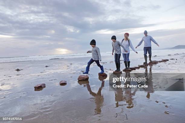 family jumping onto stepping stones on beach in winter - family trip in laws stock-fotos und bilder